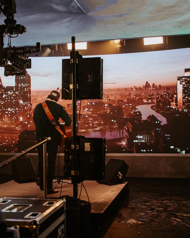 A musician performs on a stage inside a virtual production studio, surrounded by equipment, with a city skyline displayed on a curved LED wall behind them.