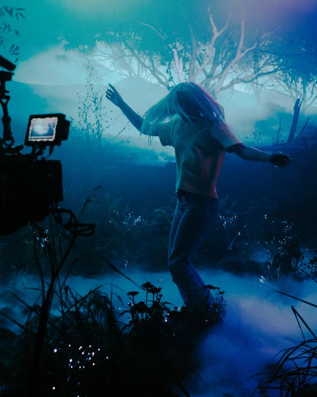 A camera operator films an actor moving through a foggy, stylized forest environment inside a virtual production stage illuminated with blue and purple lighting.