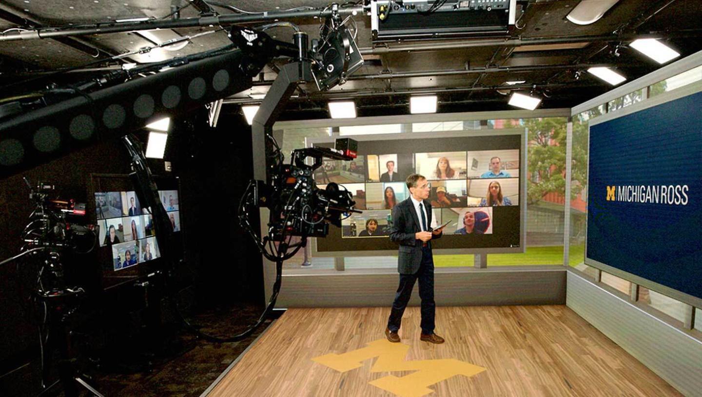 Man presenting on stage featuring people on zoom